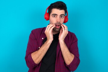 Shocked Young man standing over blue studio background stares fearful at camera keeps mouth widely opened wears wireless stereo headphones on ears