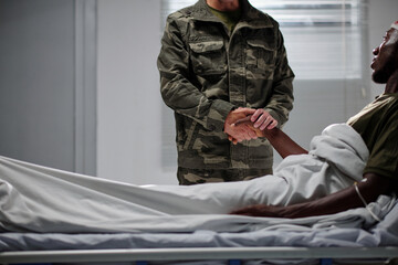 Soldier in uniform holding hand of his colleague and supporting him during his visit in hospital