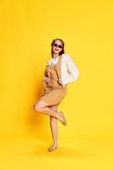 Portrait of adorable girl with long hair wearing sunglasses and dress posing at camera over yellow background
