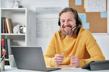 Portrait of mature teacher in headphones smiling at camera while using laptop for online lesson