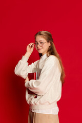 Portrait of young student girl wearing uniform, holding notebooks and looking at canera over red background