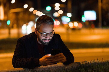 A smiling man with glasses is using a smartphone in the city with a nightlight in the background.