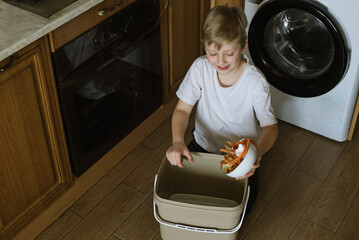 Happy Smiled Blond Boy In Kitchen Making Compost Scraping Vegetable Leftovers Into Bin at Home. Zero waste concept. Processing and recycling organic.