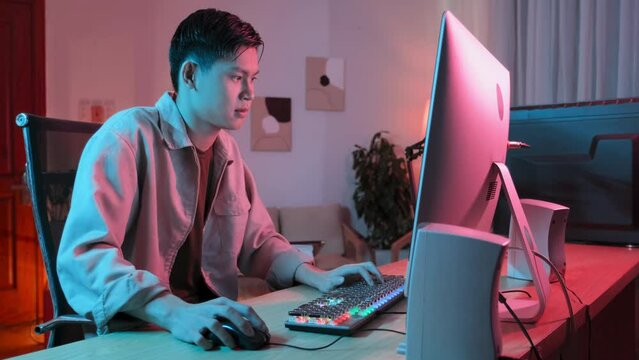 Waist up of Asian gen z gamer playing exciting online computer game in room with neon pink lighting