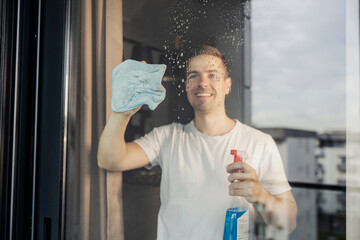 A happy tidy man is rubbing window glass at home with cloth and detergent.