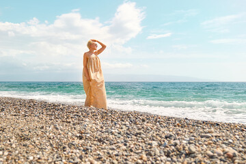Pretty blond woman walking on pebble beach enjoying sunny windy day near blue sea. Travel concept. Pastel muted color.