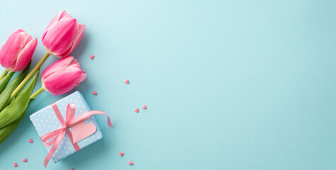 Mother's Day decorations concept. Top view photo of pink tulips small giftbox with ribbon bow and heart shaped sprinkles on isolated pastel blue background with blank space