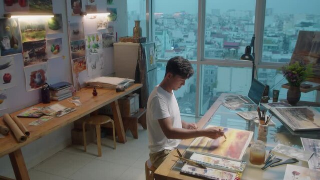 High angle wide shot of young male artist painting abstract picture with watercolors at workplace in creative studio with panoramic windows