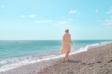 Pretty blond woman in sunglasses and transparent dress walking in the beach, smiling and enjoying...