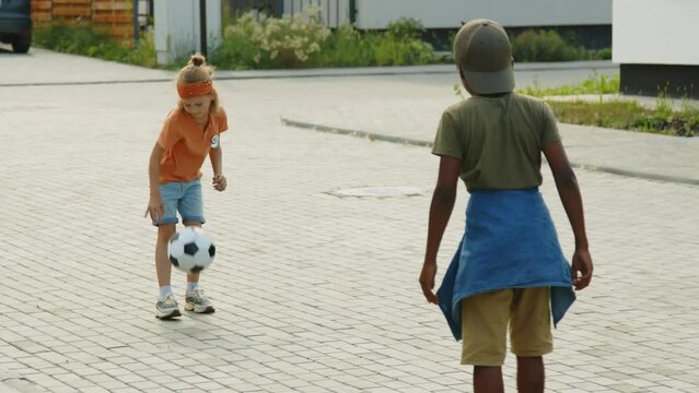 Full Shot Of Two Young Kids Hanging Out Together In Summer, Caucasian Kid Bouncing Football On Ground, Then Starting To Kick It Around With African American Friend