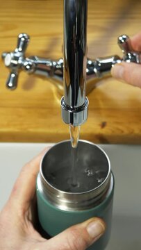 Vertical video social media format – Cinemagraph - Video of fresh running water flow from a sink tap or faucet, combined with a still image of a hand holding an insulated steel bottle as it fills up.