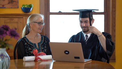 Mother and son sitting at dining room table doing online graduation ceremony in costume and holding diploma out to camera.