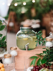 A glass lemonade dispenser sits on a decorated table at an open-air buffet. Glass tall candlesticks, ripe cherries, green leaves.