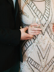 A bride in an art deco dress with heimonic beaded patterns holds a bosal of white wine. The groom in a black suit holds the bride's hand. Wedding day.