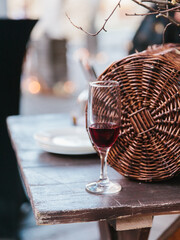 A glass of red wine stands on an old wooden table. In the background are tree branches and a wicker basket. Rural style.