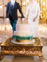 A two-tiered wedding cake with gold and green decor stands on a gold stand. In the background, the newlyweds hold hands.