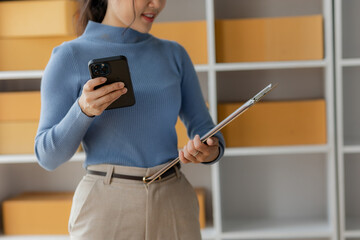 Starting an SME, an independent Asian female small business entrepreneur using a laptop and a smart phone. Cheerful success, cute smiley face. Asian woman raising her hand, packaging box, online marke