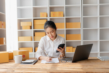 Starting an SME, an independent Asian female small business entrepreneur using a laptop and a smart phone. Cheerful success, cute smiley face. Asian woman raising her hand, packaging box, online marke