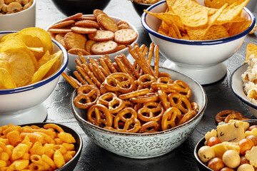 Salty snacks, party mix. An assortment of crispy appetizers in bowls on a table. Potato and tortilla chips, crackers, popcorn etc - obrazy, fototapety, plakaty