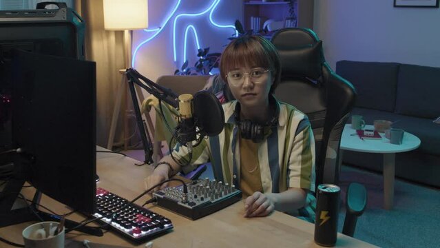 Medium portrait shot of professional Chinese female gamer sitting alone in her home studio with blue neon lighting, with computer, soundboard, energy drink on desk, and looking straight at camera