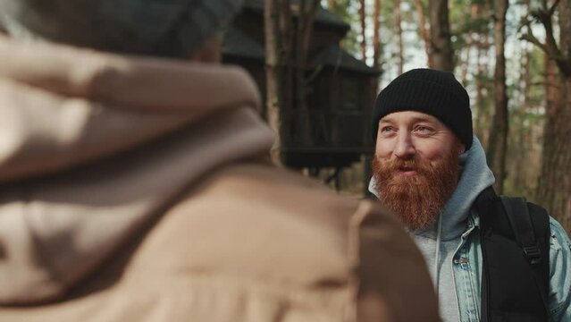 Positive bearded redheaded hiker talking with his friend in the woods with a backpack