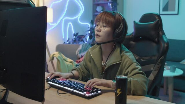 Waist-up shot of nerdy young Asian woman wearing glasses and headset sitting in her room with blue neon light, playing intensive videogame on computer and sipping energy drink