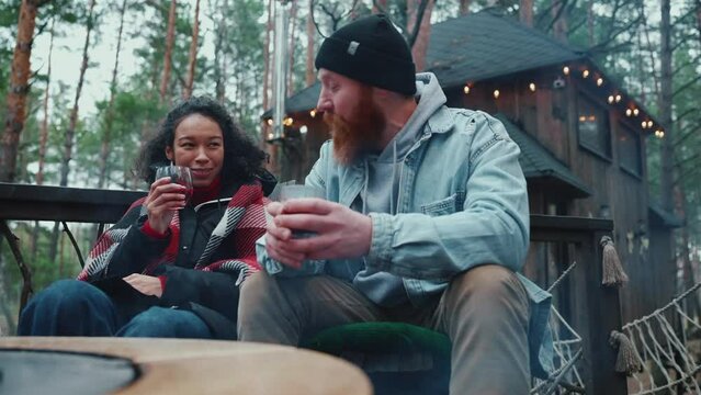 Multinational couple talking and drinking mulled wine around a campfire in the forest