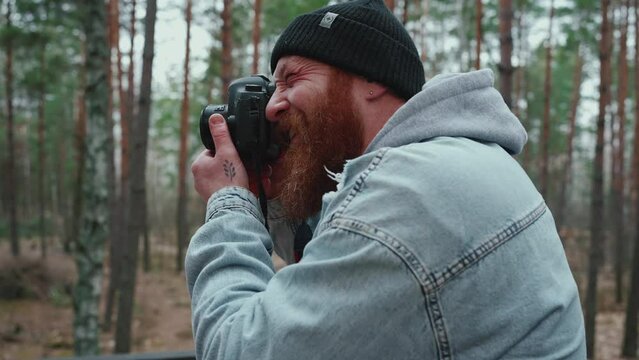 Cheerful red haired man taking pictures on his camera in the forest