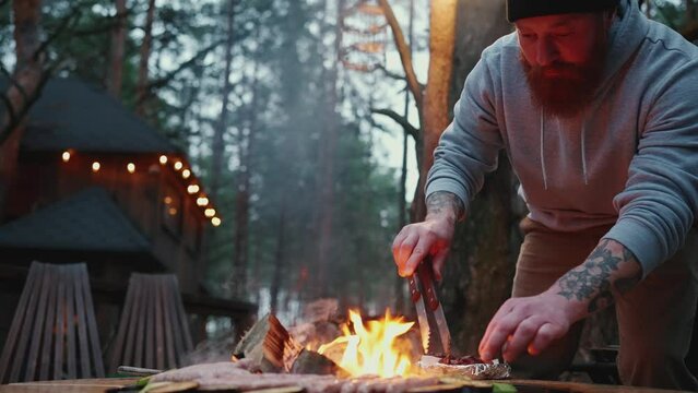 Confident bearded man frying sausages with zucchini on the fire in the forest