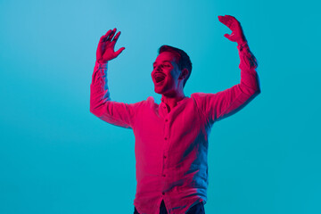 Fototapeta na wymiar Portrait of young emotional man raising hands with positive happy expression, posing against blue studio background in pink neon light. Concept of youth, emotions, facial expression, lifestyle. Ad