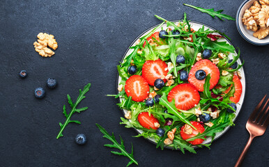 Strawberry and herbs healthy salad with arugula, blueberries and walnuts, black kitchen table, place for text. Fresh useful vegan dish for healthy eating