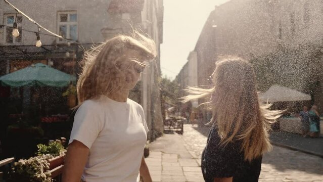 Camera follows two beautiful happy young female friends in round sunglasses walk through street spray, smiling at camera