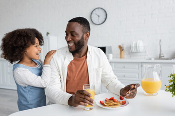 happy african american girl looking at cheerful father having breakfast in kitchen.