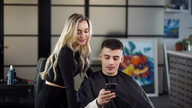 A woman hairdresser and a male client in a beauty salon look at photo references for cutting and styling hair on a smartphone