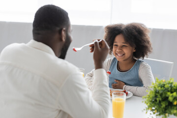 happy african american girl having breakfast and looking at father eating strawberry on blurred foreground.