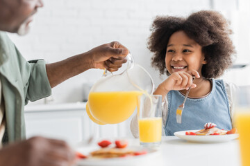 happy african american girl looking at grandfather pouring orange juice during breakfast.