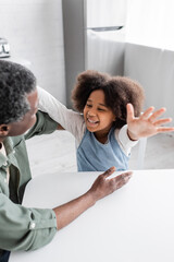 happy african american kid with curly hair smiling while stretching hands towards grandfather at home.