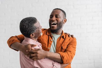 Positive african american son hugging mature father in living room.