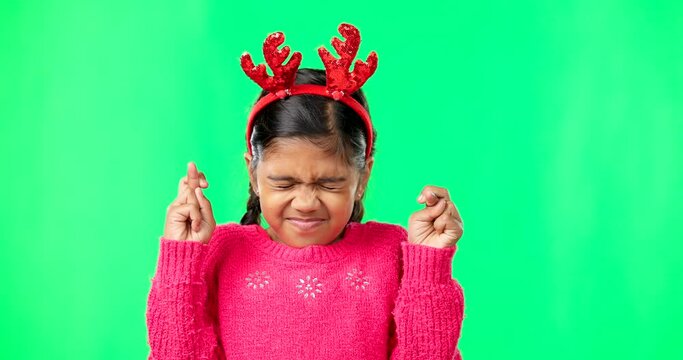 Child, christmas and wish on green screen fingers crossed and antlers for luck. Girl kid or eyes closed emoji for hope, praying and miracle or faith on studio background space with reindeer headband