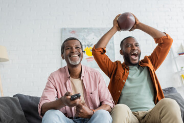 Cheerful african american man holding remote controller near son with rugby ball at home.