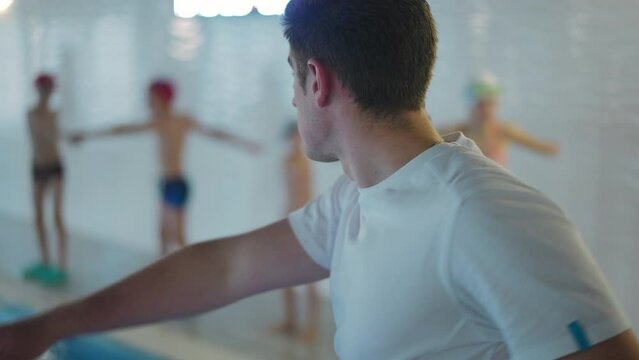 Shooting over shoulder of trainer training group of blurred unrecognizable children in swimming pool. Back view confident Caucasian young man exercising with students indoors