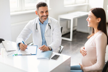 Prenatal care concept. Happy young pregnant lady sitting at clinic, having consulation with male gynecologist
