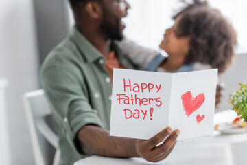 african american man holding greeting card with happy fathers day lettering near curly daughter on blurred background.