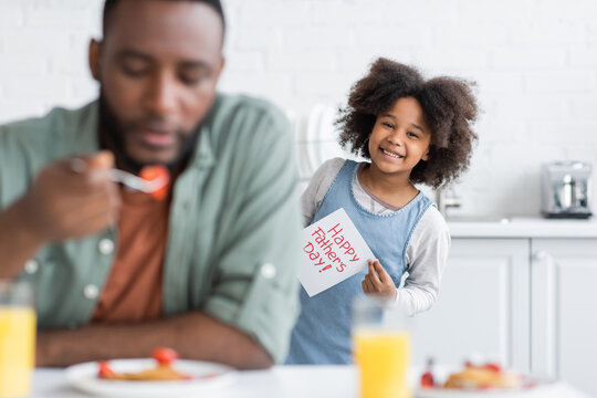 cheerful african american girl holding greeting card with happy fathers day lettering while standing behind dad on blurred foreground.