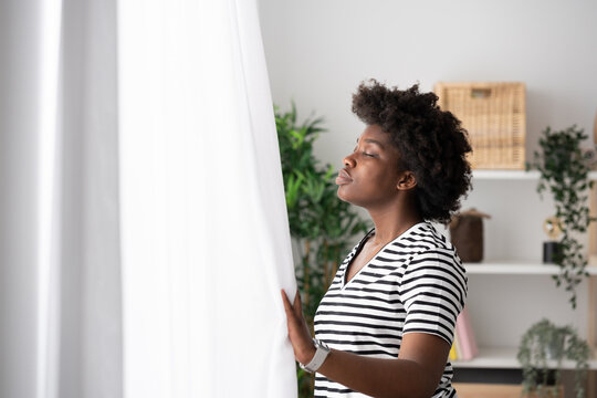 Young African American woman breathing deep at home beside window with closed eyes.