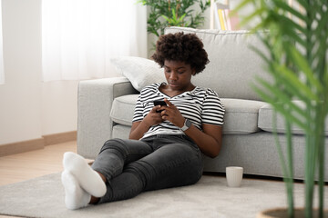 African American woman using phone sitting on the floor at home. Internet and social media concept