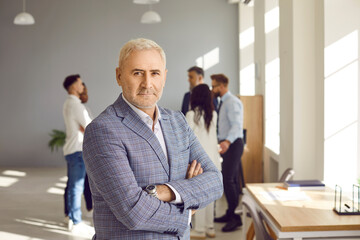 Portrait of mature senior confident business man leader in suit proudly looking at the camera with crossed arms and with a team of company employees talking in background in the office.