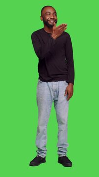 Vertical video: Front view of african american man sending air kisses on camera, posing with kissy face on full body greenscreen. Young adult being romantic in studio and doing flirty gesture