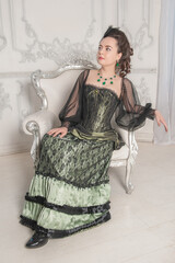 Beautiful young woman wearing green medieval vintage Victorian Style dress sitting in the armchair