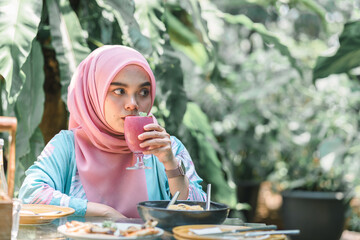 Cheerful and beautiful Muslim Asian woman in a pink hijab drinking a berry fruit smoothie at an outdoor restaurant.